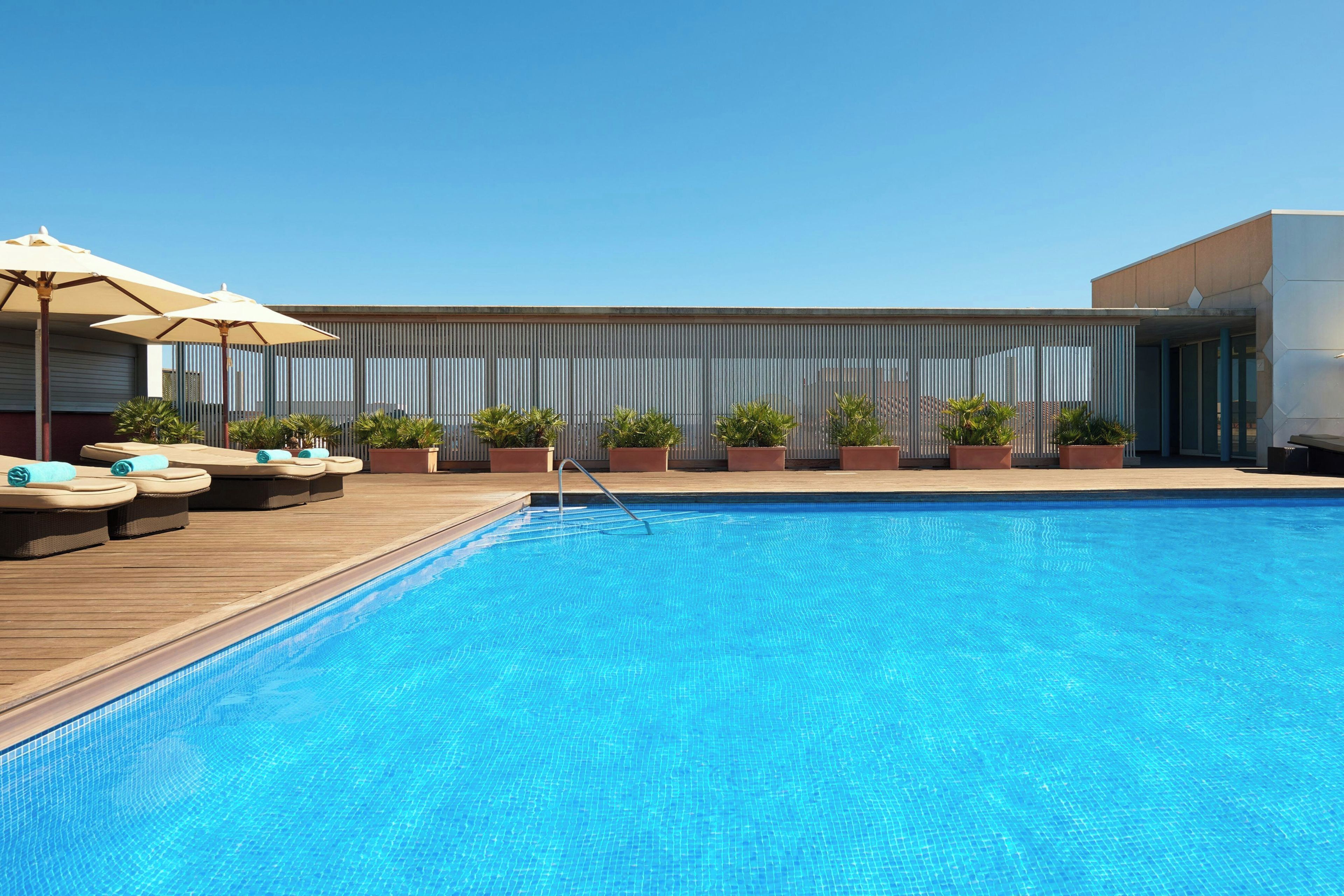 A swimming pool with lounge chairs and umbrellas, surrounded by potted plants and a modern building facade.