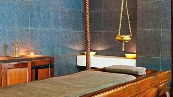 Una habitación con una cama de madera, una pequeña mesa de madera con una lámpara, y un estante en la pared con un cuenco.