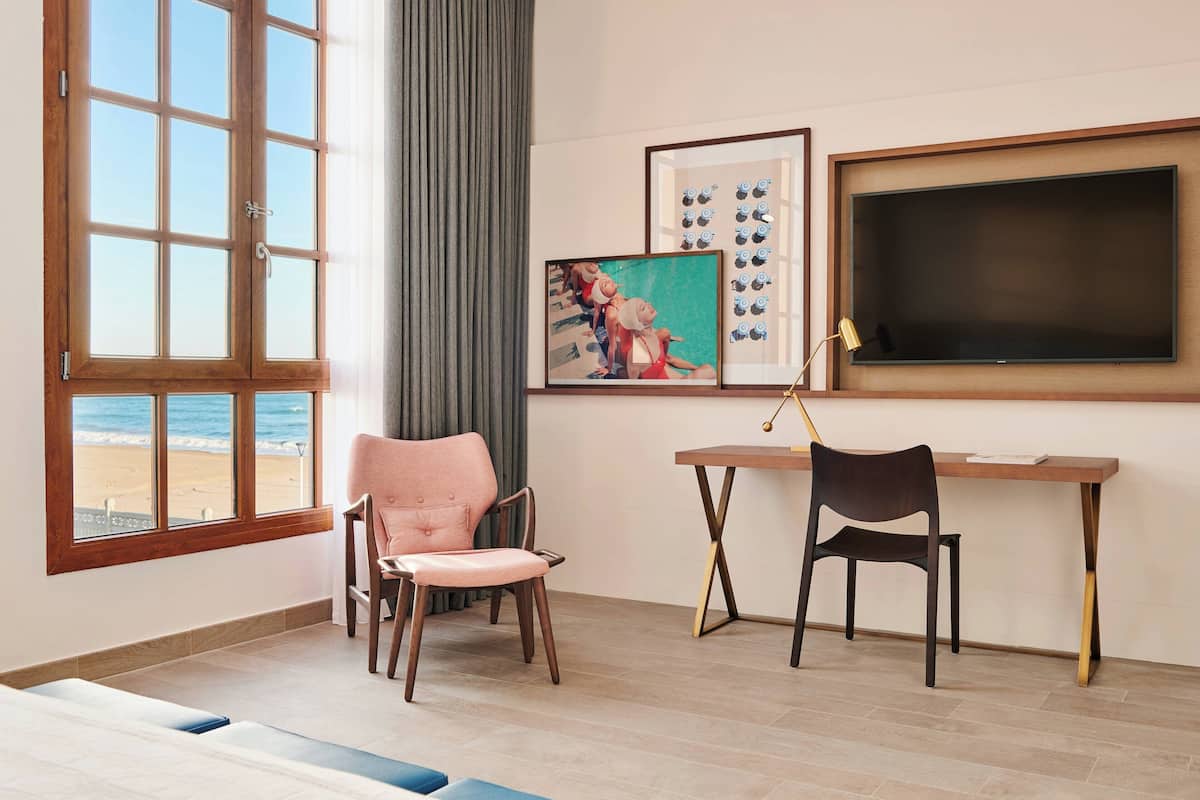 A room with a large window overlooking a beach, a desk with two chairs, and a framed picture on the wall.