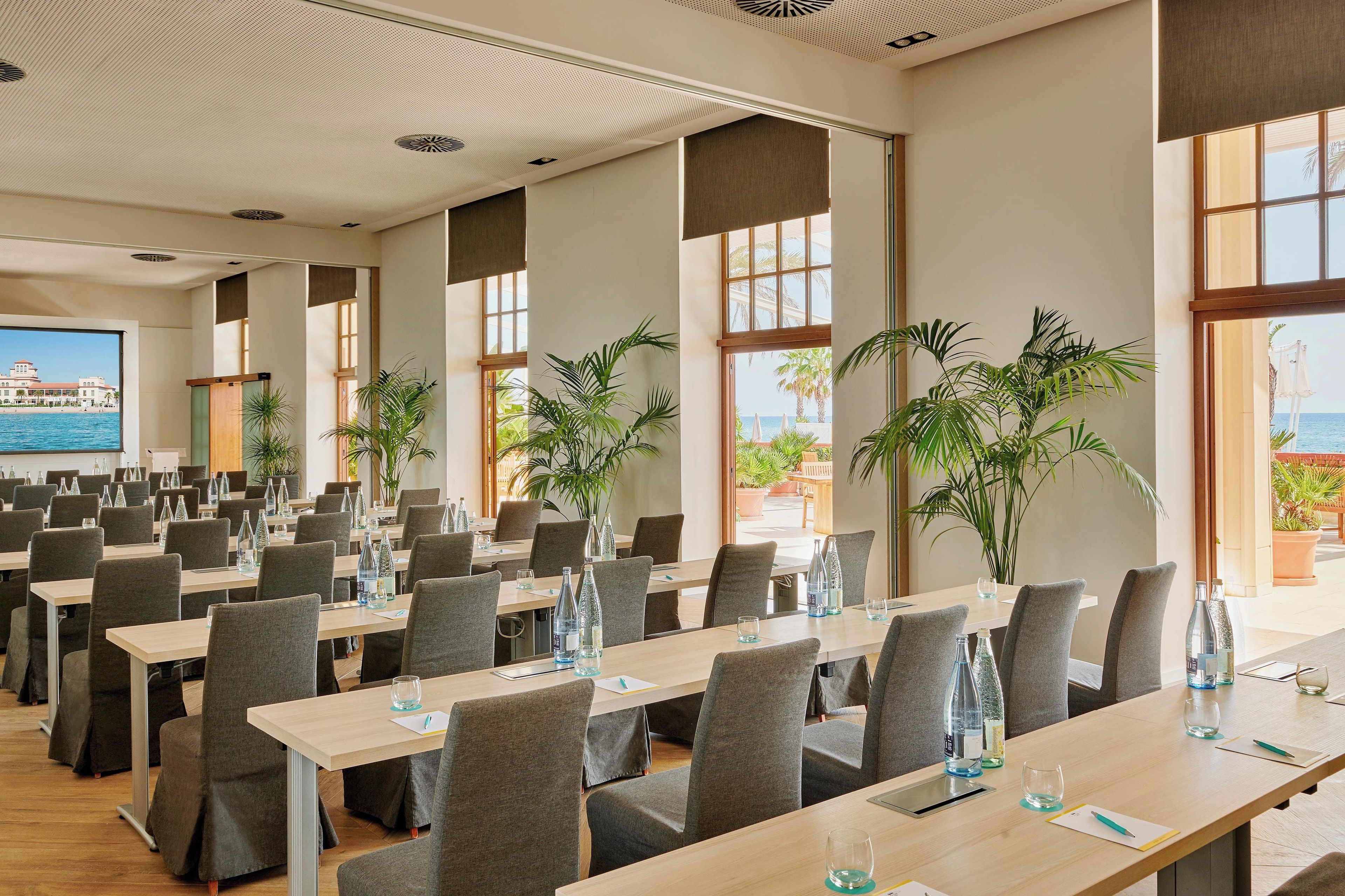 A conference room with multiple tables and chairs, large windows with views of the sea, and potted plants.