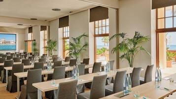 A conference room with multiple tables and chairs, large windows with views of the sea, and potted plants.