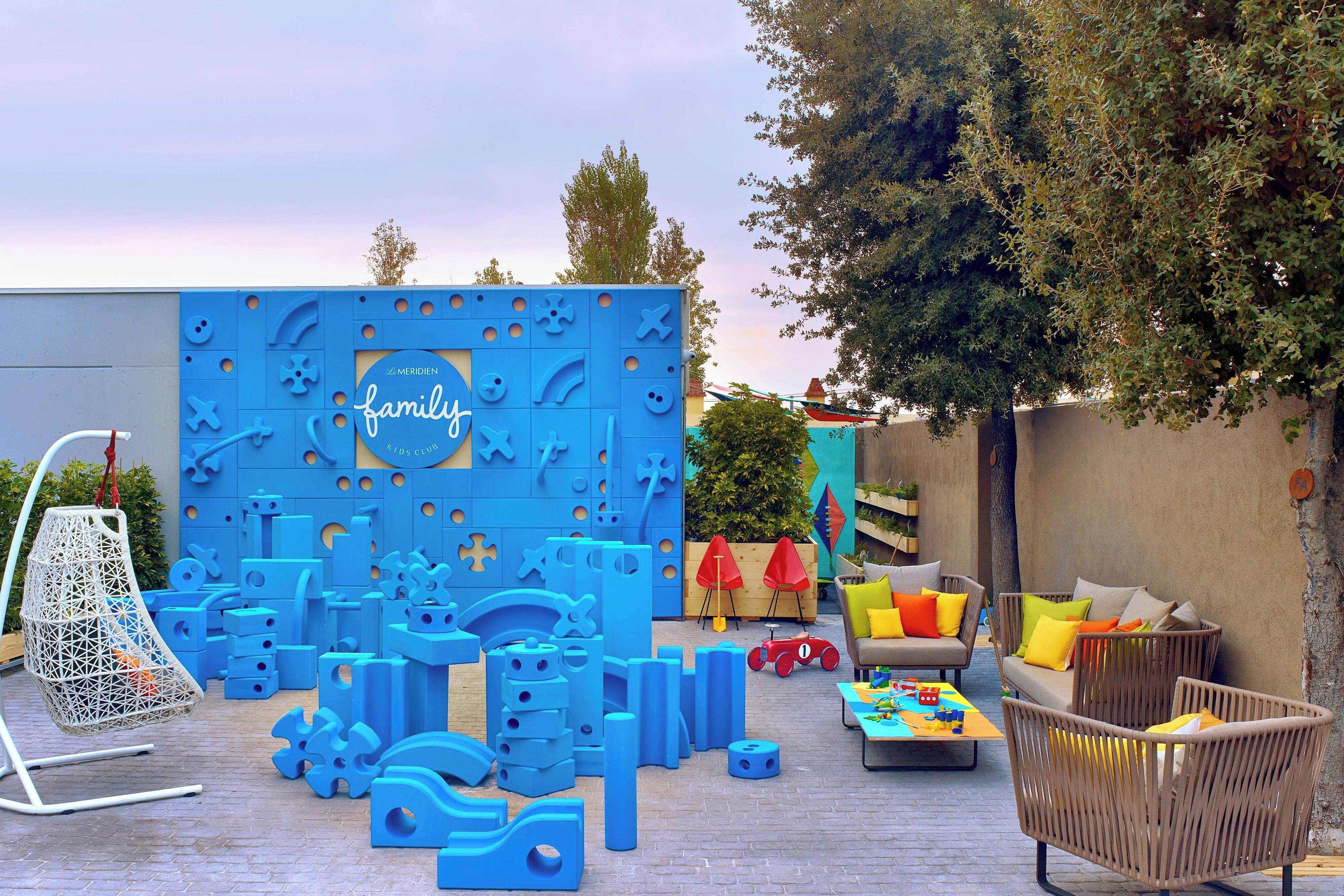 Un espace de jeux pour enfants avec des structures bleues, une chaise suspendue et des sièges d’extérieur dotés de coussins.