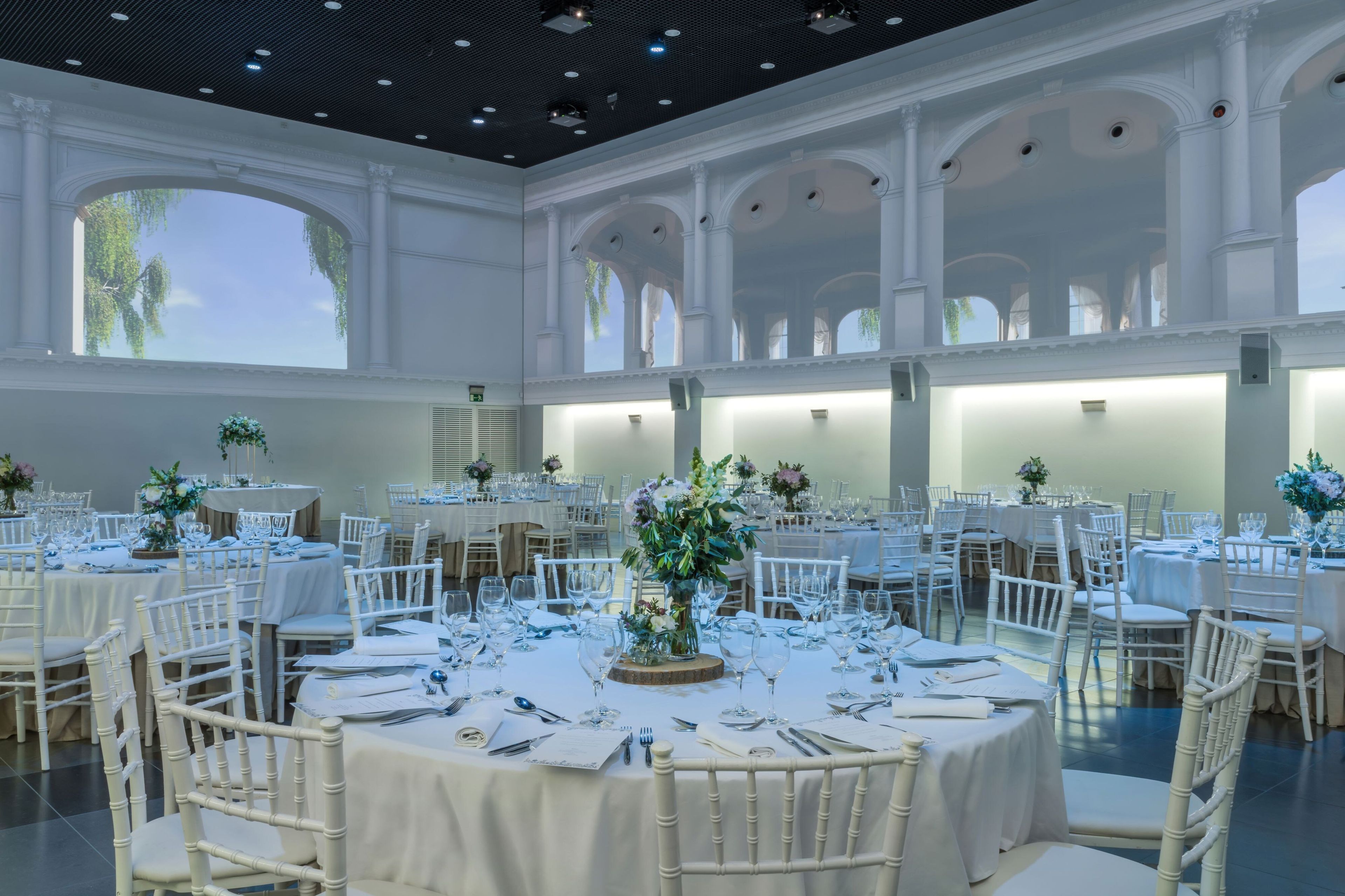 Uma ampla sala de banquetes com várias mesas redondas, cada uma com toalhas brancas, cadeiras e centros de mesa.