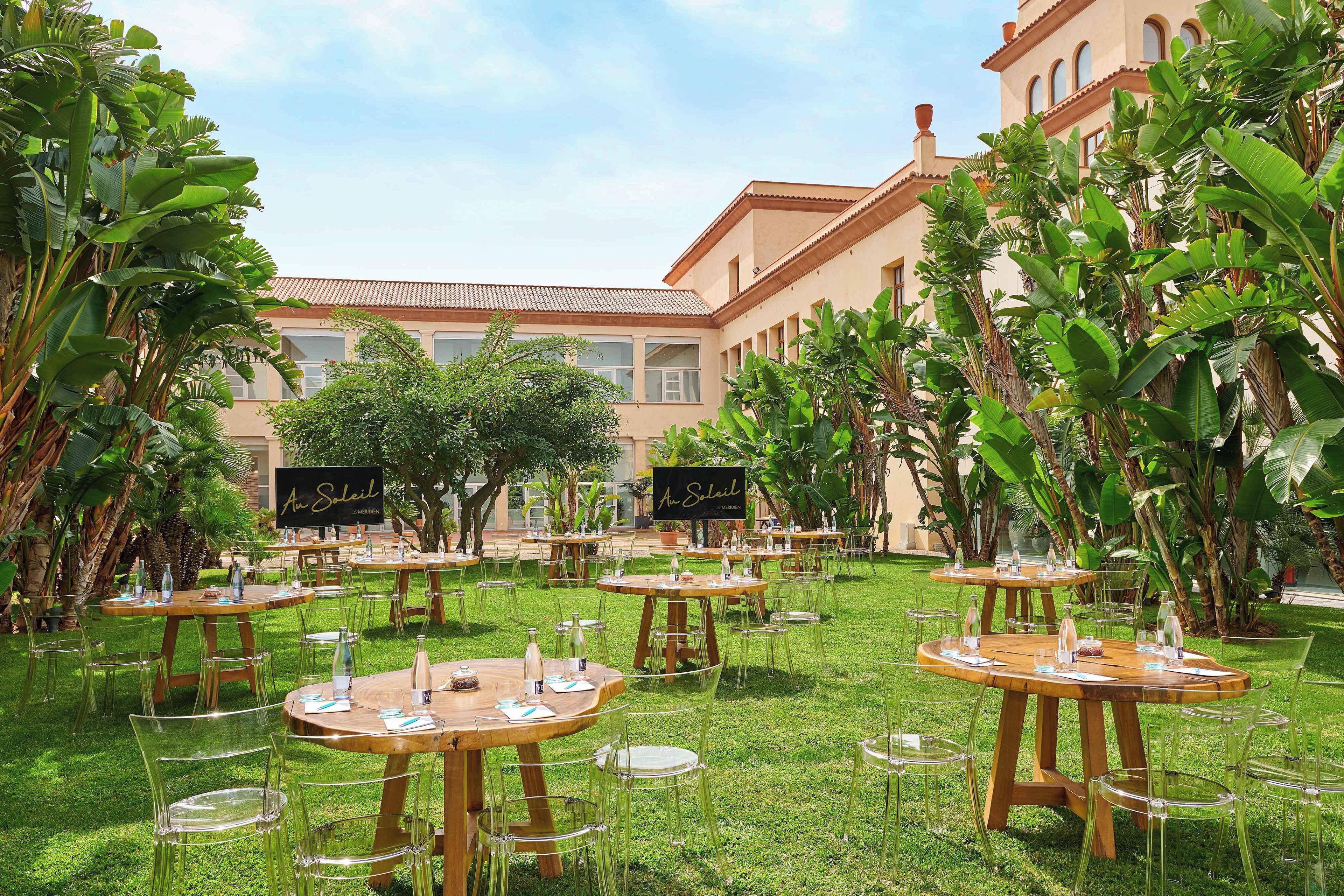 Un área de comedor al aire libre con mesas de madera redondas y sillas transparentes, rodeada de abundante follaje y un edificio con ventanas arqueadas.