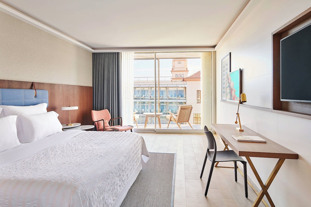A hotel room with a large bed, a desk, a chair, a window with a view of buildings, and a balcony.