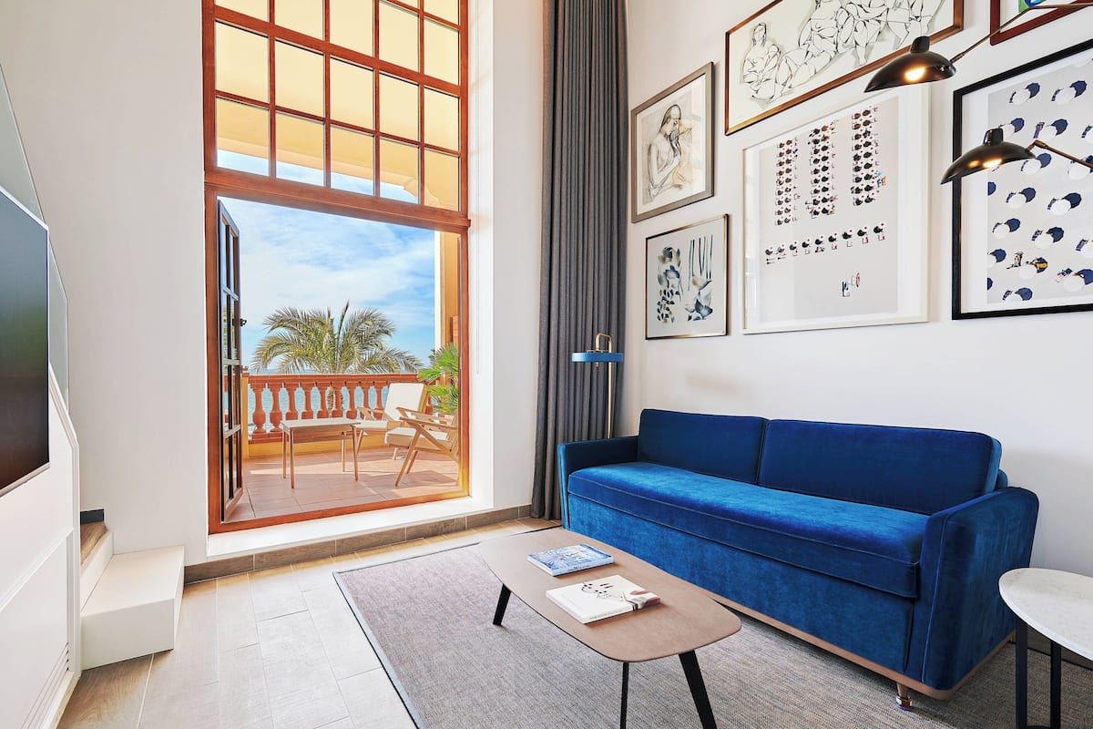 A living room with a blue sofa, a coffee table, and a TV. There are framed artworks on the wall and a large window with a view of palm trees.