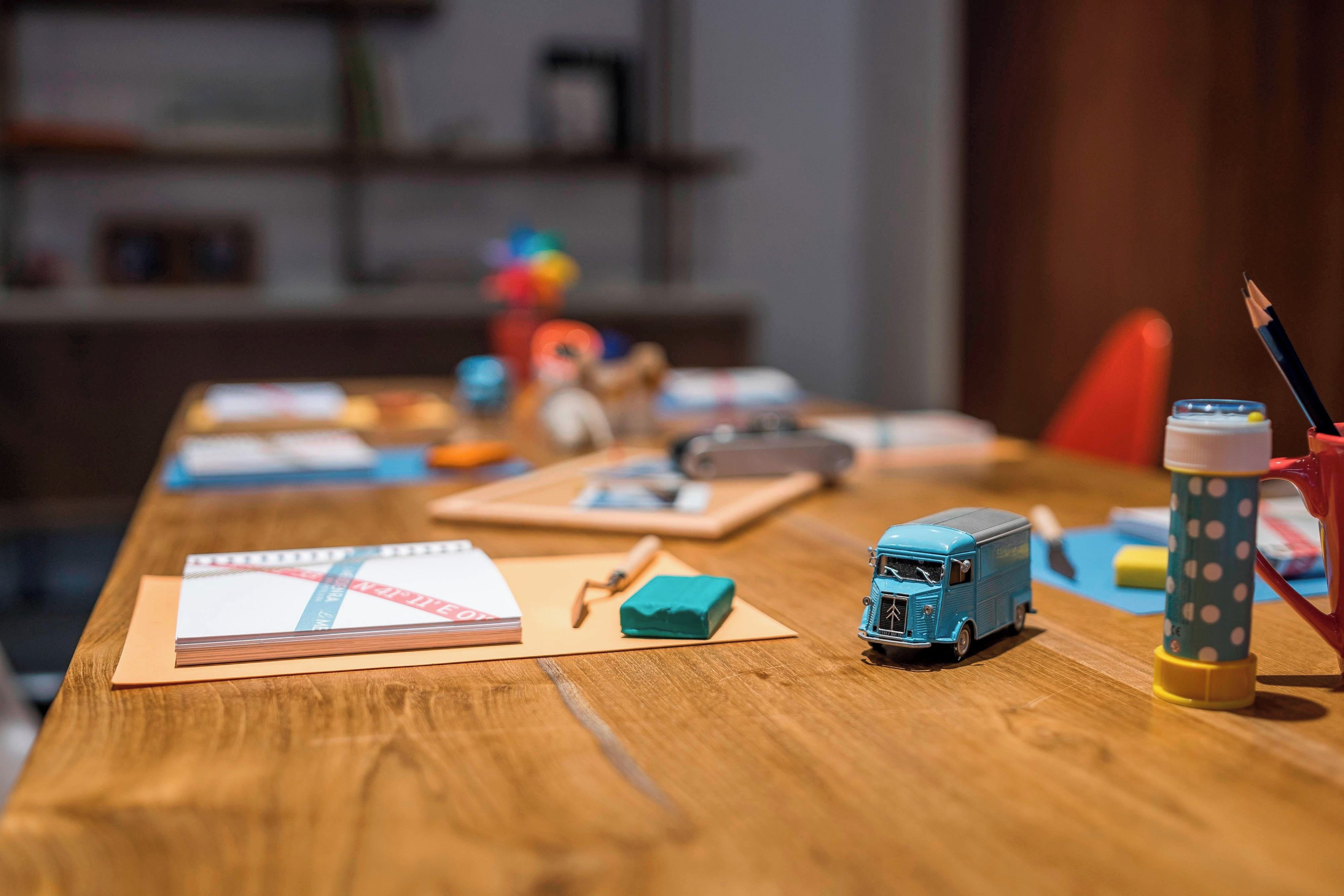 A wooden table with a notebook, a small blue toy bus, a pencil, and a polka-dotted cup.