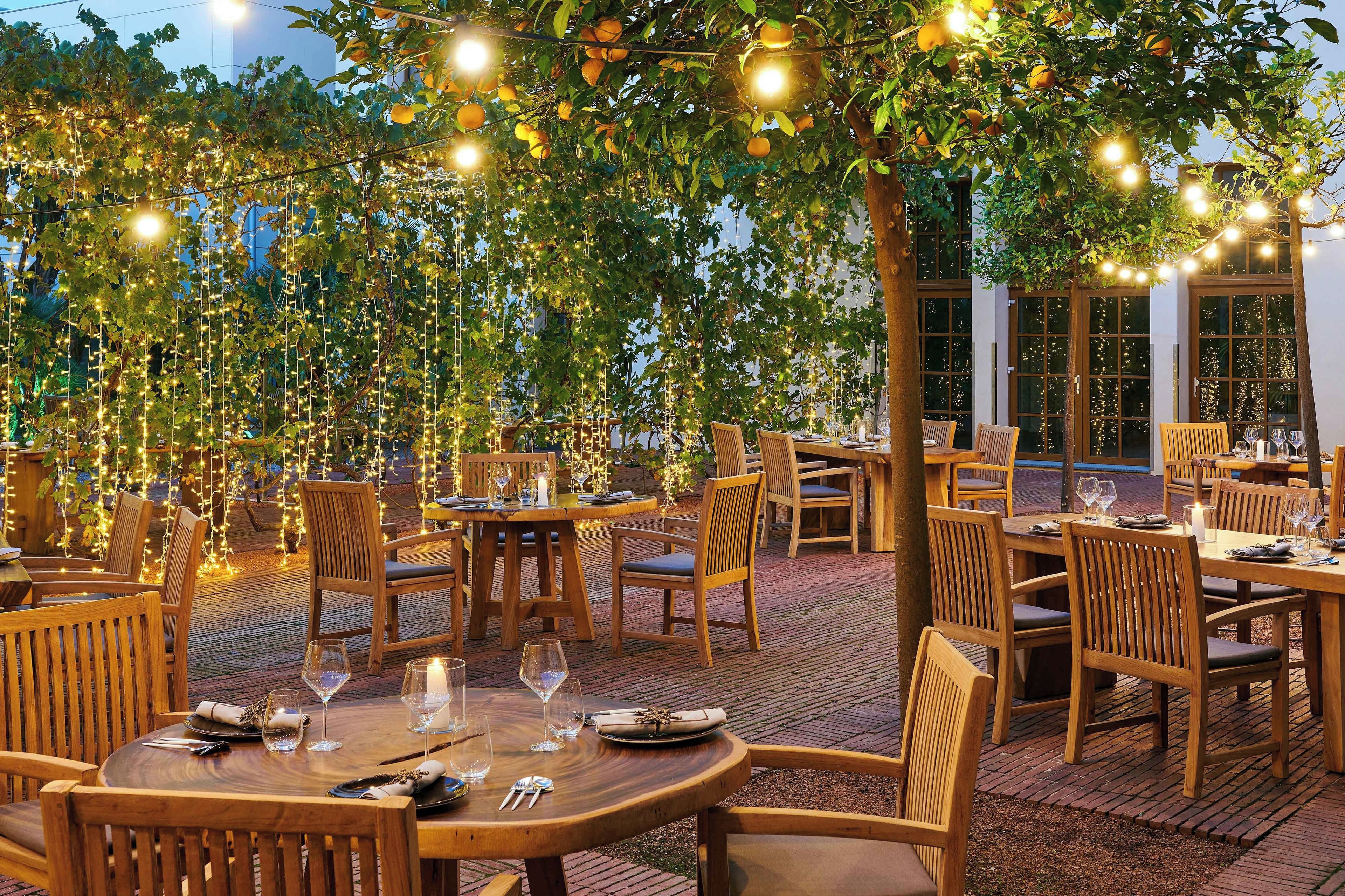 Un espace extérieur aménagé pour les repas, avec des tables et des chaises, des guirlandes lumineuses et un sol en briques.
