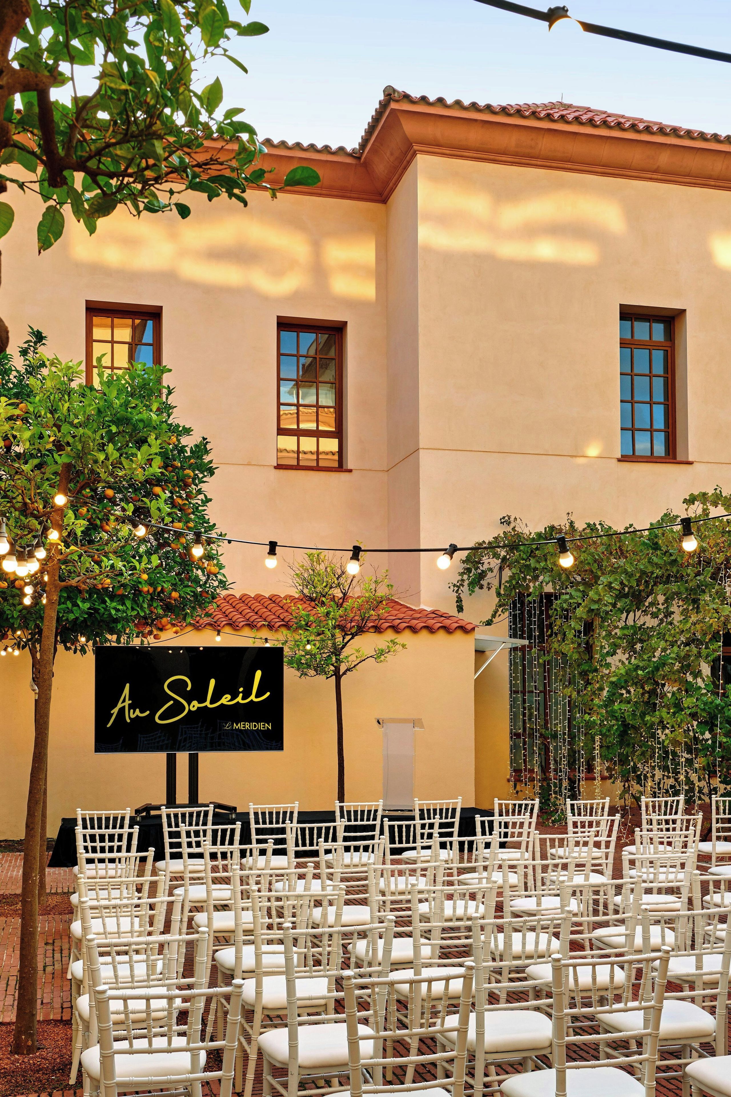 A building with a tiled roof, a sign reading "An Soleil", and rows of white chairs arranged for an event.
