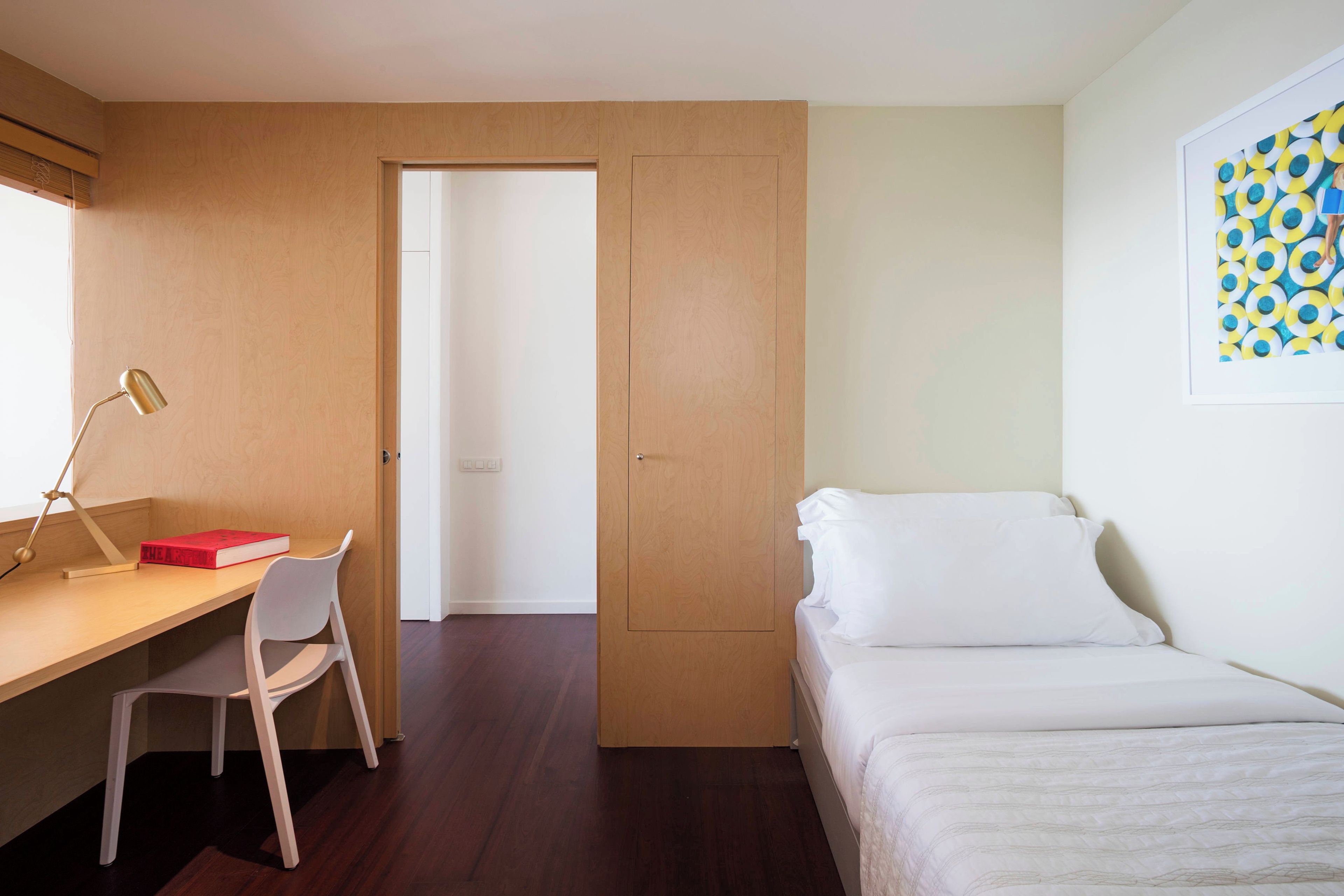 A room with a desk, chair, bed, and a door leading to another room.