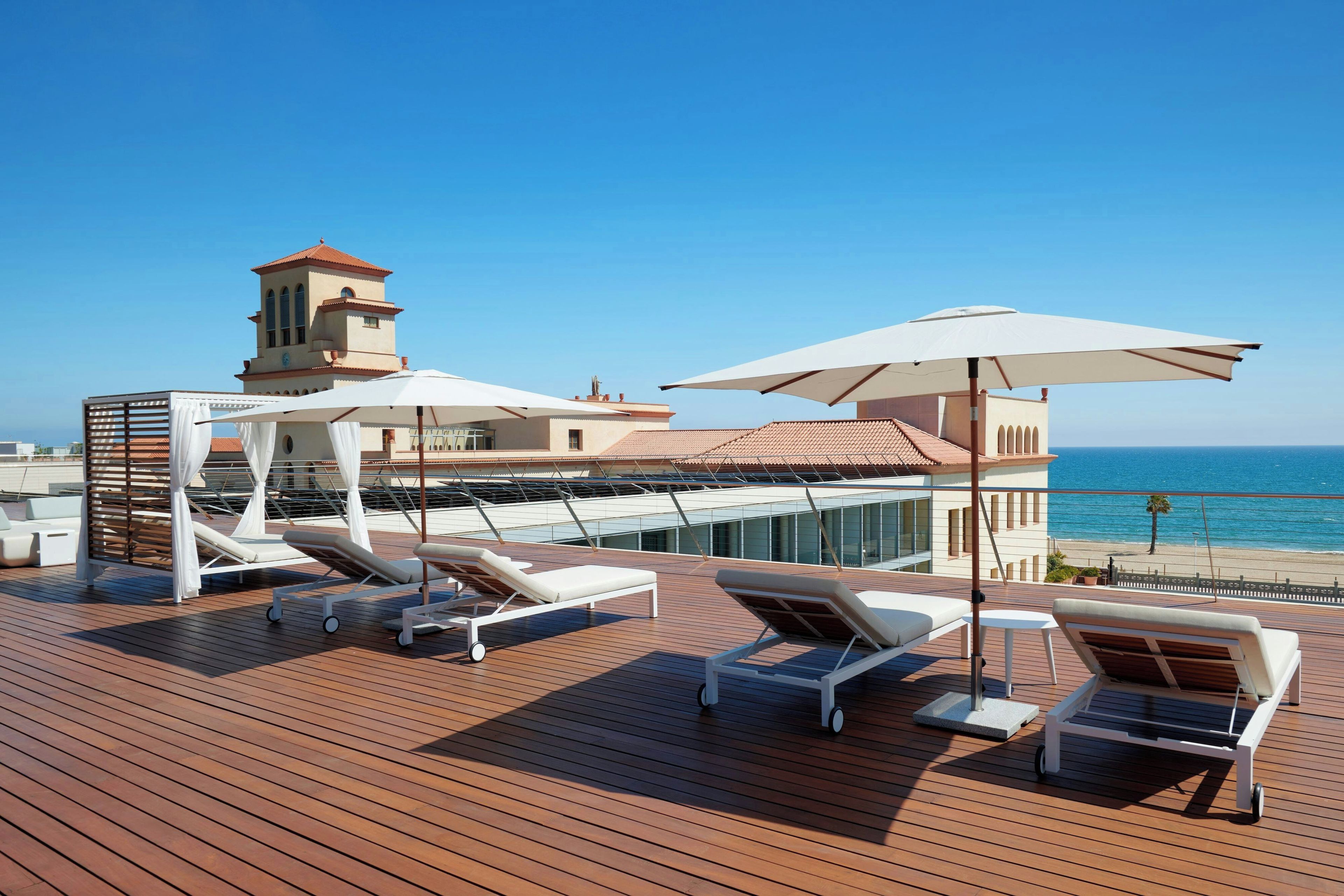 A wooden deck with sun loungers and umbrellas, overlooking a beach and a building with a tower.