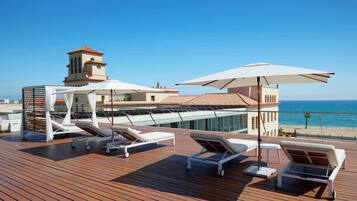 A wooden deck with sun loungers and umbrellas, overlooking a beach and a building with a tower.