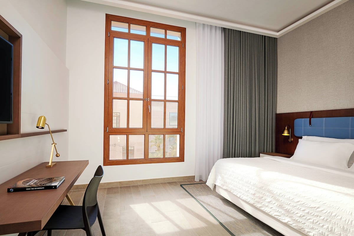 A hotel room with a large window, a desk with a lamp, a bed with white bedding, and a blue headboard.