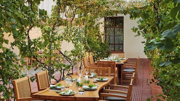 Un área de comedor al aire libre con mesas y sillas de madera, rodeada de follaje y luces colgantes.