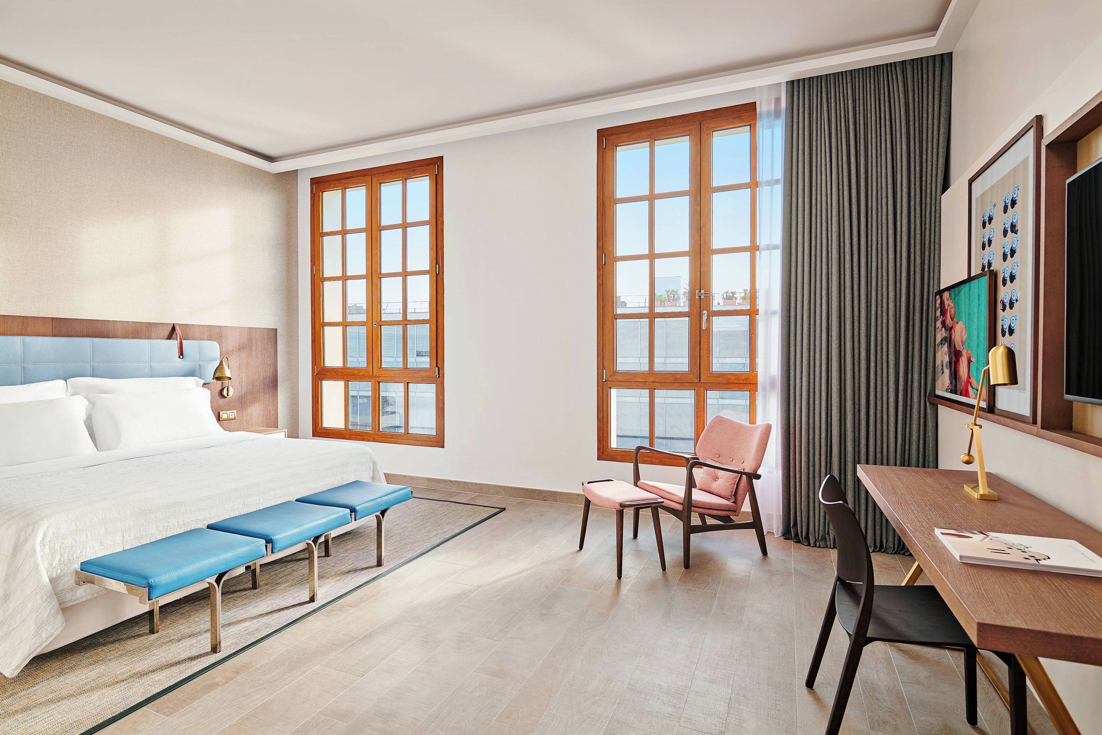 Una habitación de hotel con cama, escritorio, silla, banco y ventanas amplias.