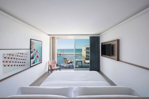 Una habitación de hotel con una cama grande, un televisor, un cuadro enmarcado y vistas al mar a través de una puerta corrediza.