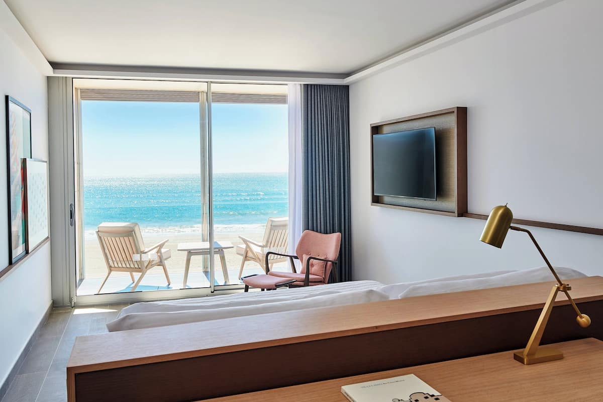 A hotel room with a large sliding glass door leading to a beach view, a bed, a desk with a lamp, and a TV on the wall.