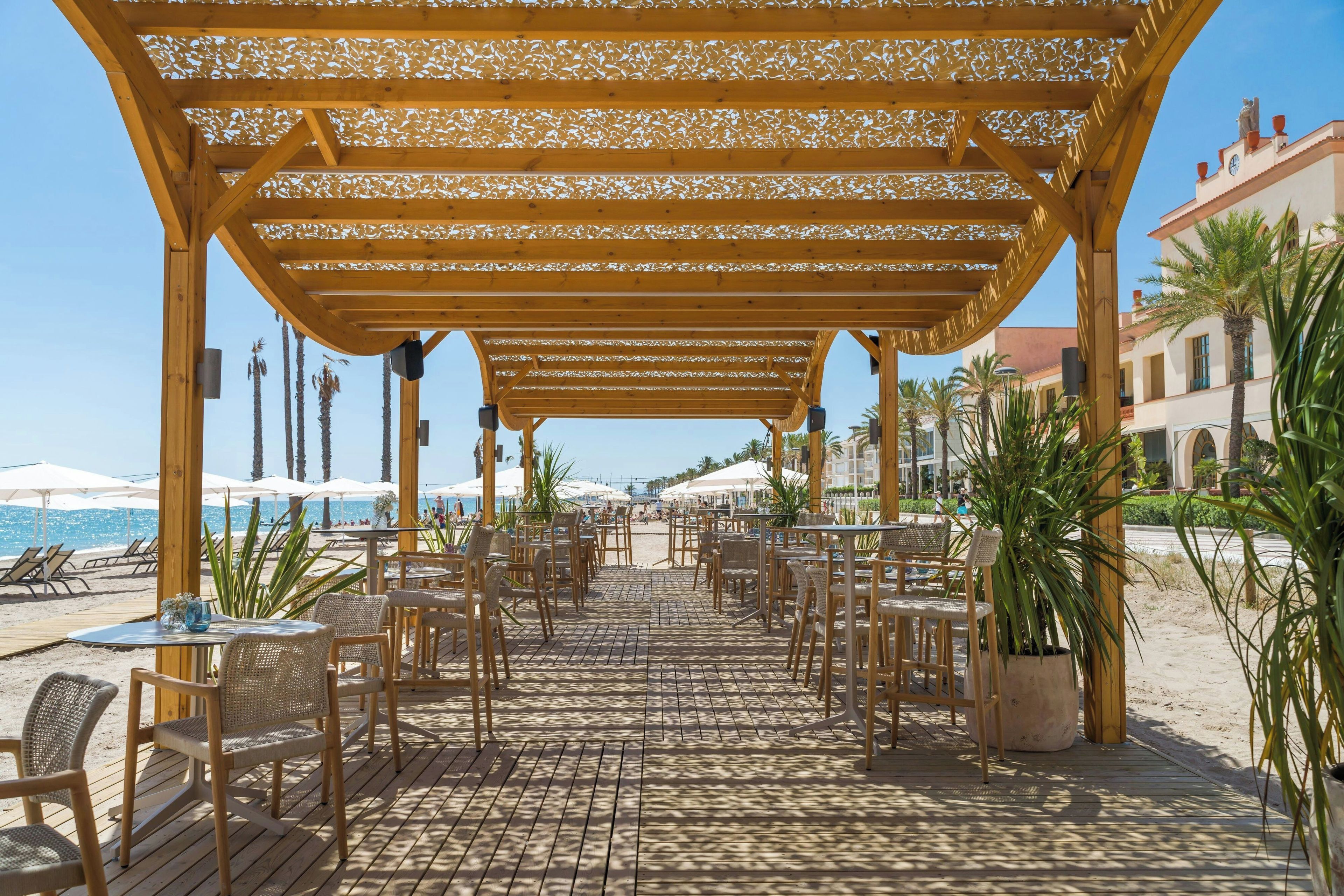 An outdoor patio with wooden tables and chairs, a pergola overhead, and a view of the beach and ocean.