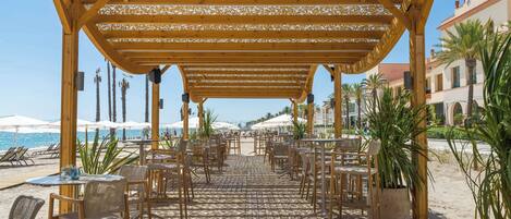 Un patio al aire libre con mesas y sillas de madera, una pérgola sobre la cabeza y vistas a la playa y al océano.