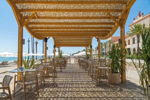 An outdoor patio with wooden tables and chairs, a pergola overhead, and a view of the beach and ocean.