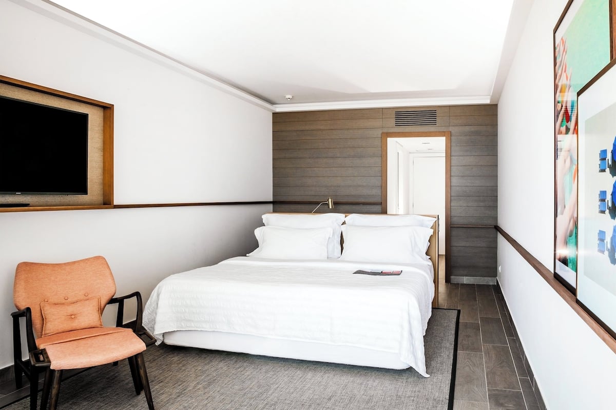 A hotel room with a bed, a chair, a TV, and a door leading to another room.