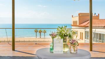 Terraza de madera con una mesa que sostiene un menú y arreglos florales, con vista a una playa y el océano.