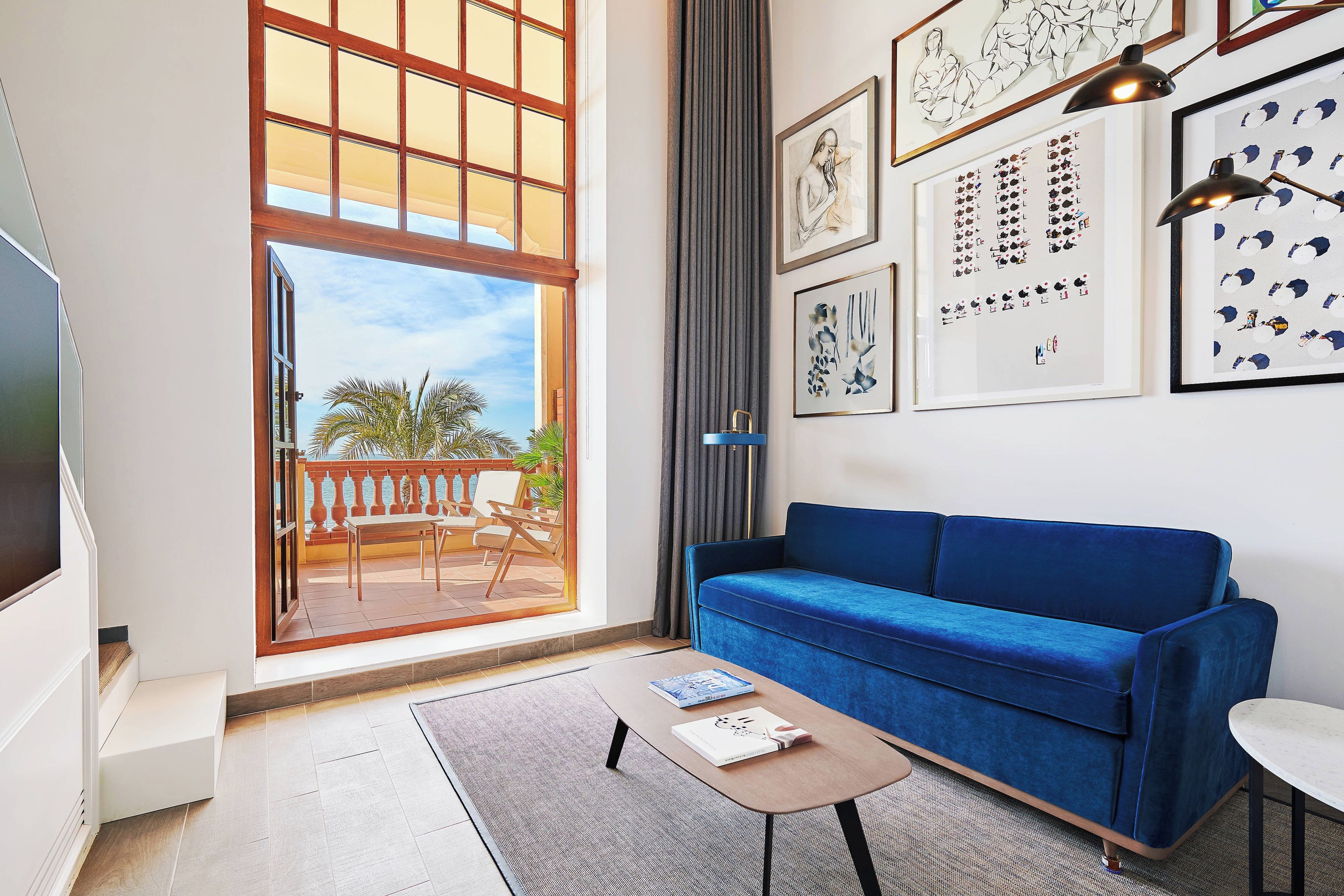 A living room with a blue sofa, a coffee table, and a TV. There are framed artworks on the wall and a view of the outdoors through a large window.