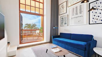 A living room with a blue sofa, a coffee table, and a TV. There are framed artworks on the wall and a view of the outdoors through a large window.