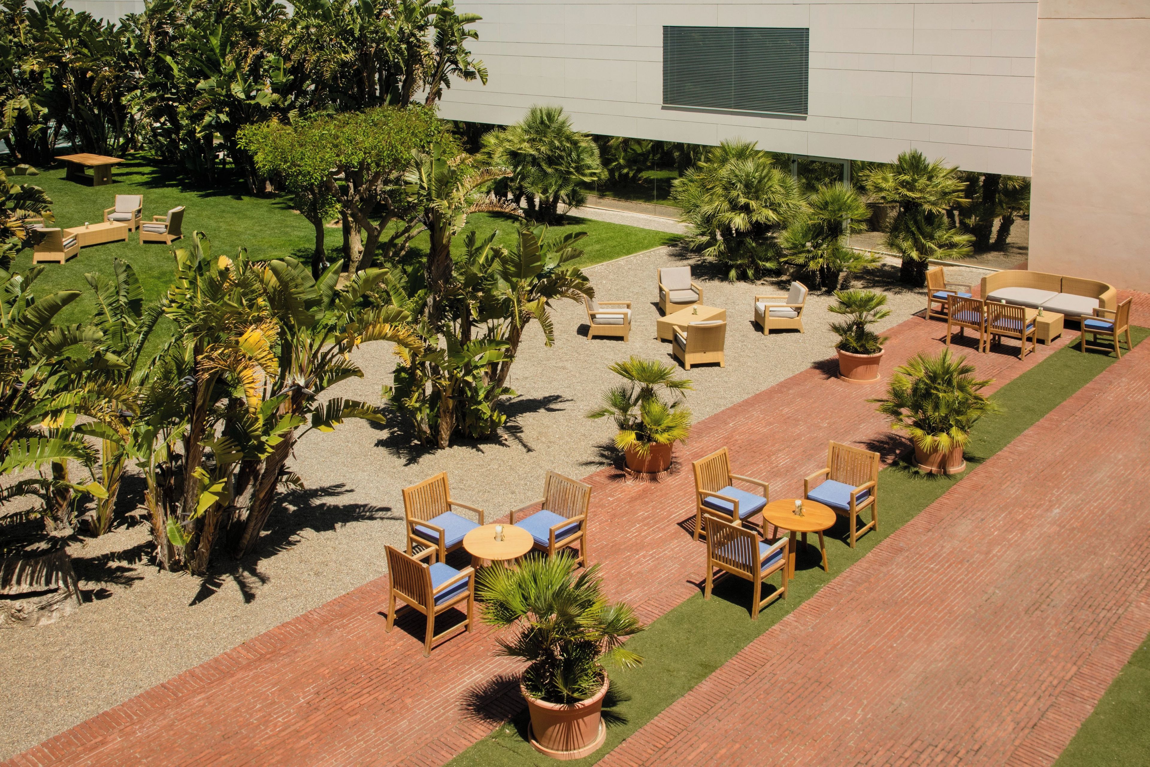 An outdoor patio area with multiple seating arrangements, including round tables and chairs, surrounded by lush greenery and palm trees.