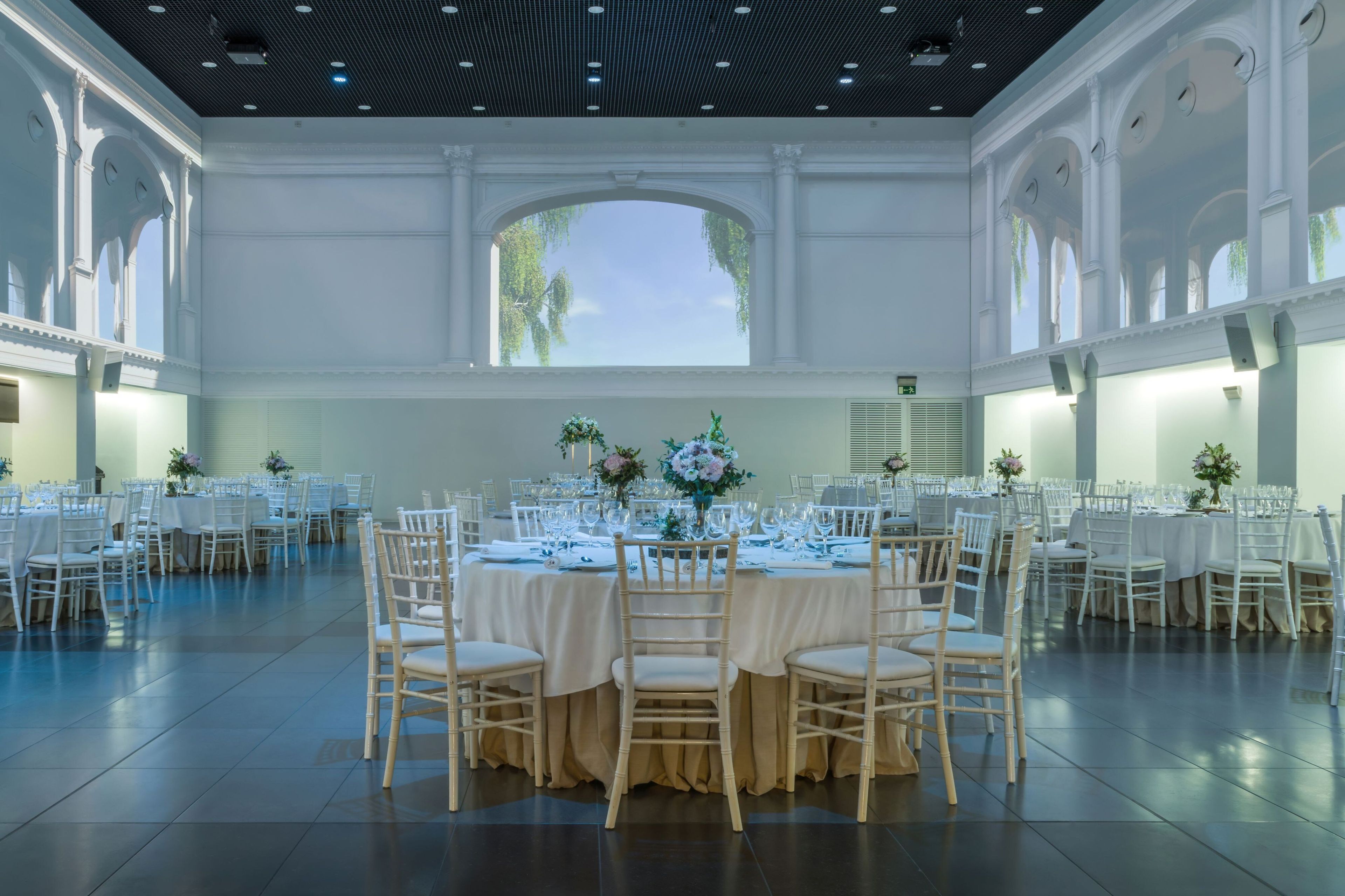 Un amplio salón de banquetes con varias mesas redondas, cada una con mantel blanco y sillas, y grandes ventanales que dejan entrar luz natural.