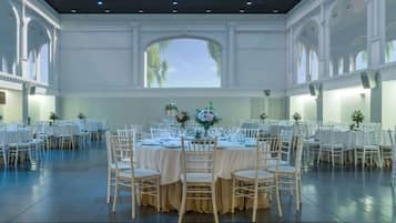 Un amplio salón de banquetes con varias mesas redondas, cada una con mantel blanco y sillas, y grandes ventanales que dejan entrar luz natural.