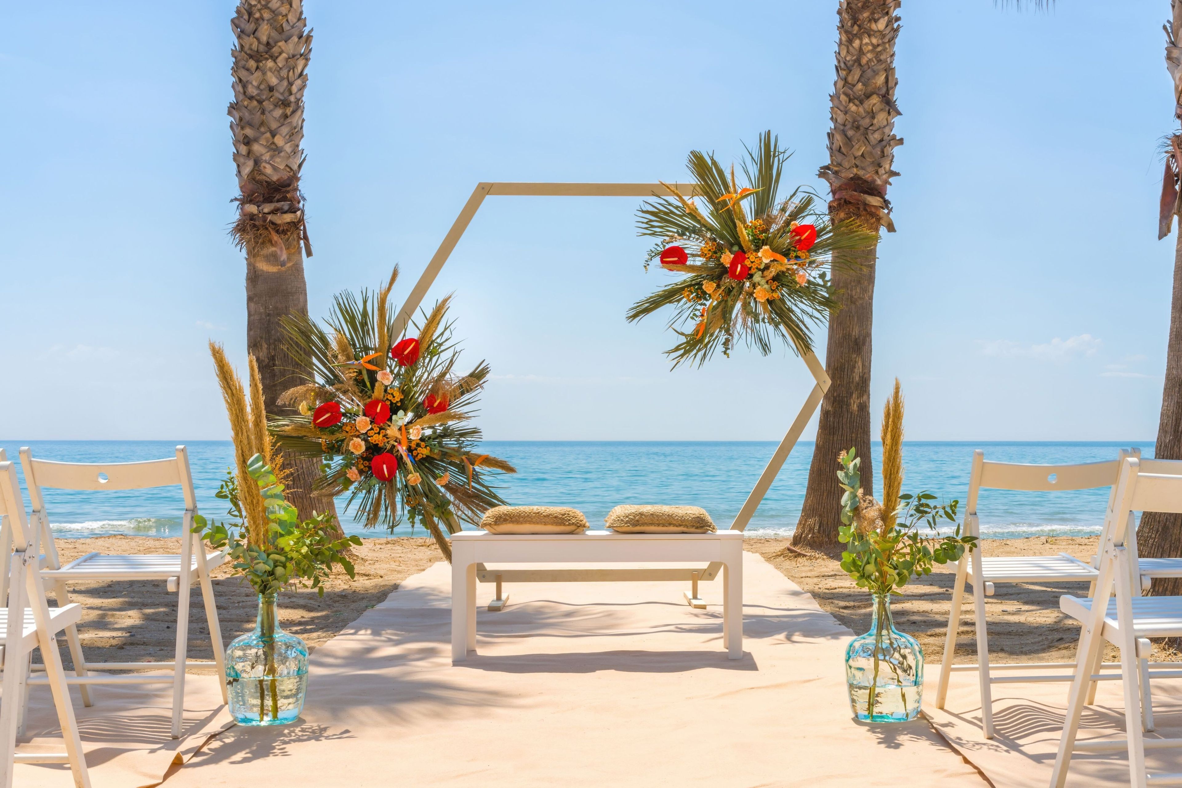 A beach wedding setup with a wooden arch, white chairs, and a bench.