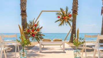 A beach wedding setup with a wooden arch, white chairs, and a bench.