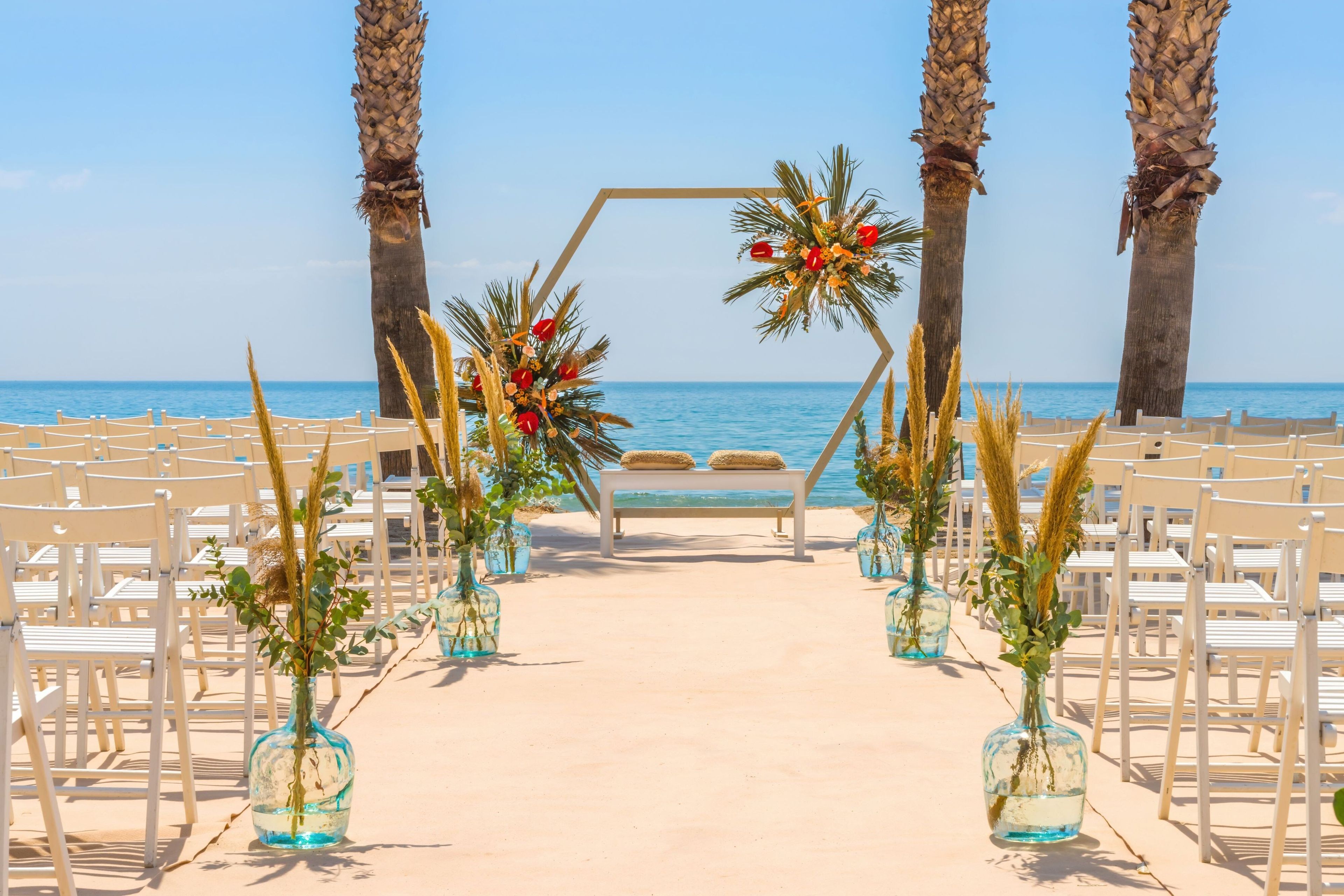 A beach wedding setup with a clear view of the sea, arranged chairs, and palm trees.