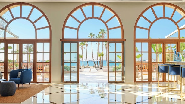 Un vestíbulo espacioso con amplias ventanas arqueadas, piso de mármol pulido y vista a una playa y palmeras.