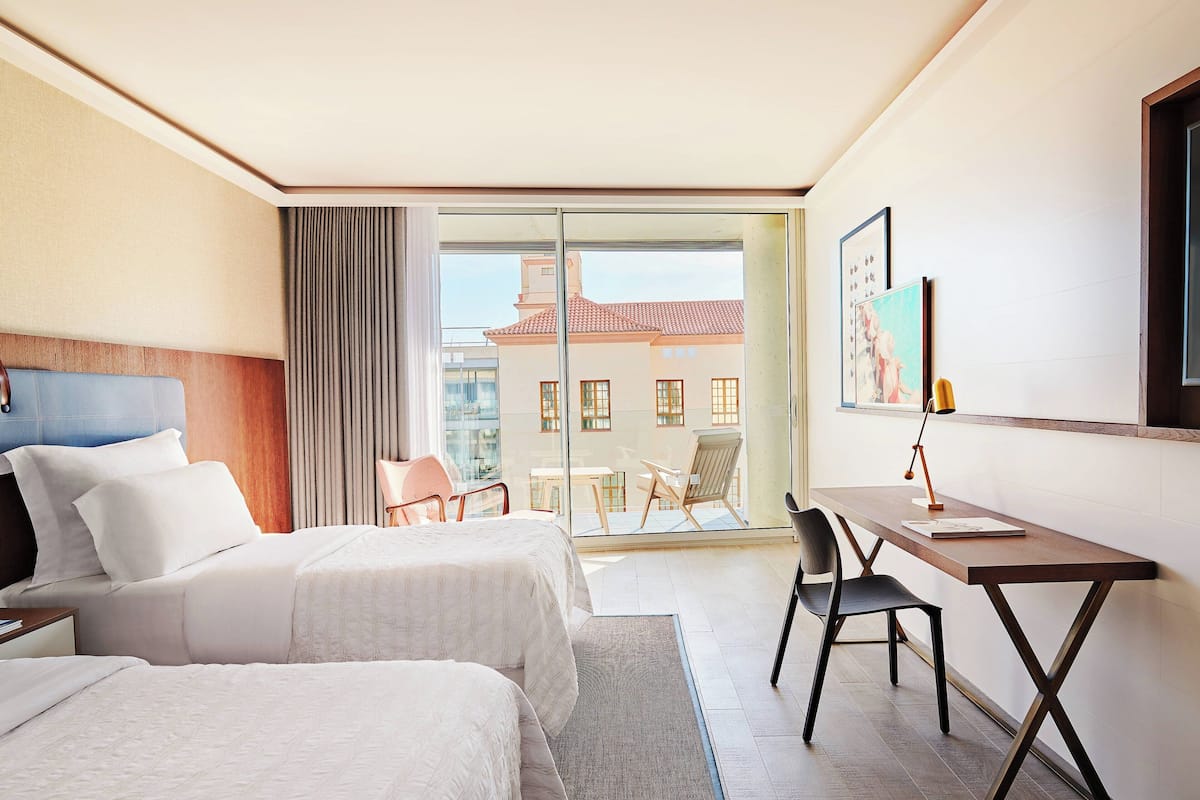 A hotel room with two beds, a desk, a chair, a large window with a view of buildings, and a painting on the wall.