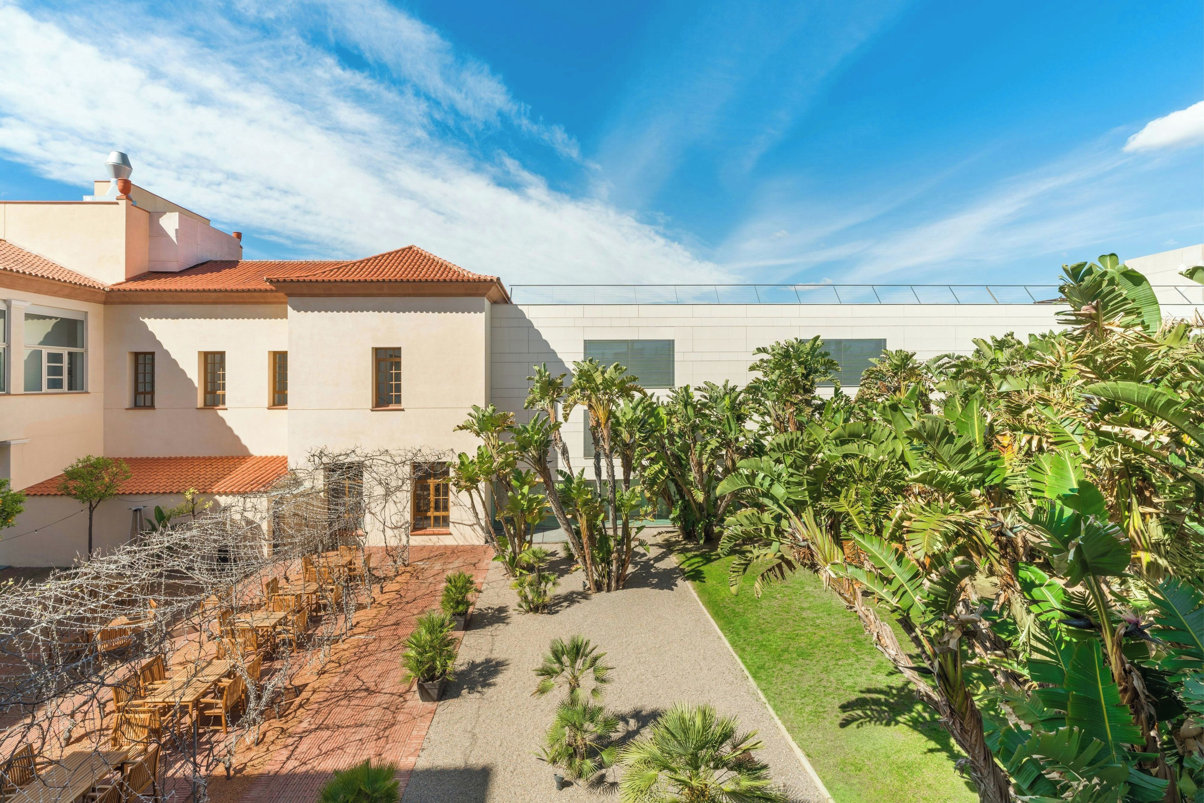 Un patio con mesas al aire libre, un edificio blanco de techo rojo y exuberante vegetación tropical.