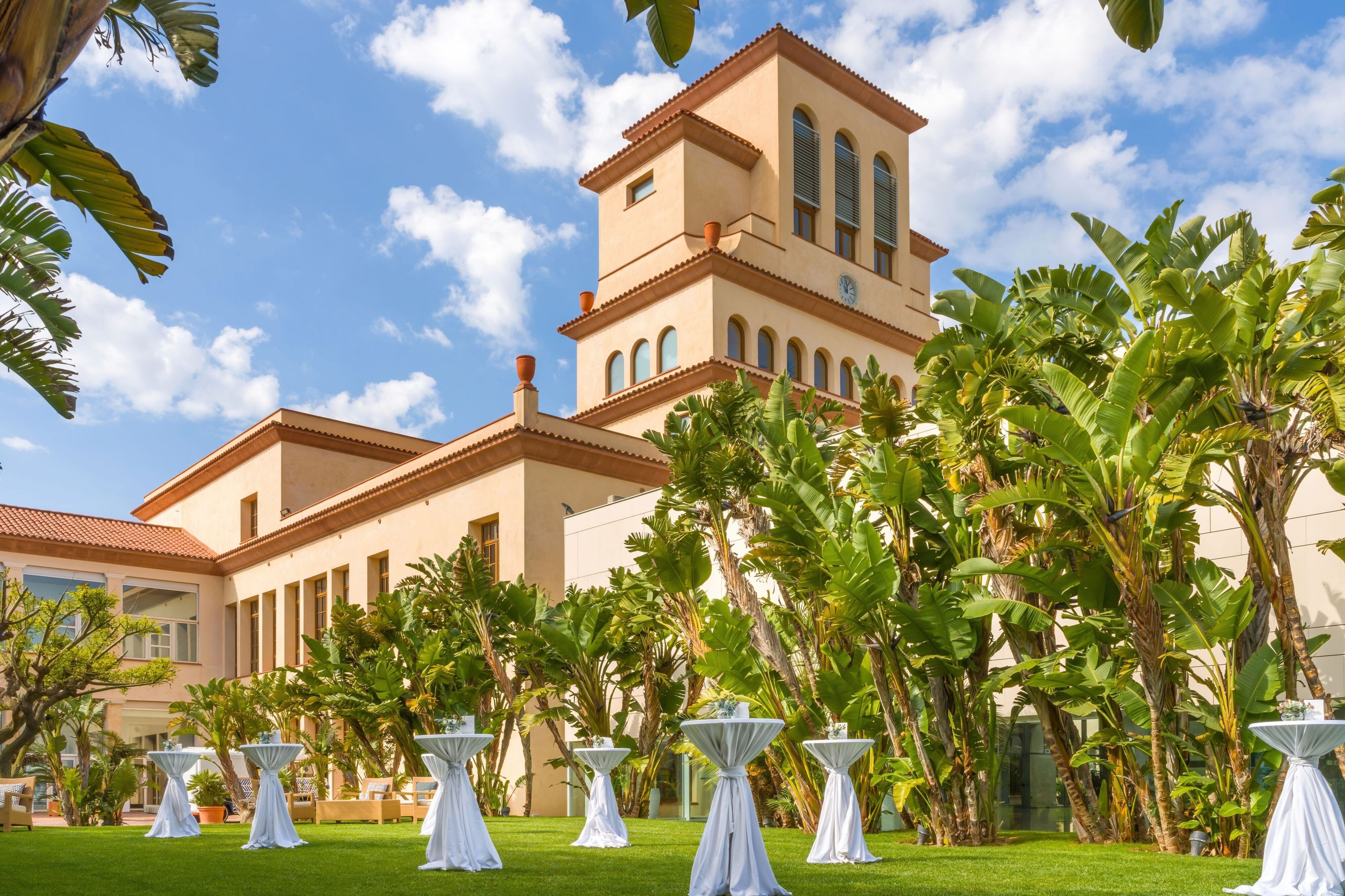 Un hotel con un edificio grande que cuenta con una torre de reloj, rodeado de exuberante follaje y mesas blancas dispuestas para comer al aire libre.