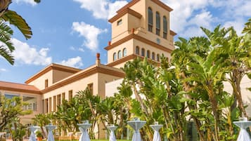 Un hotel con un edificio grande que cuenta con una torre de reloj, rodeado de exuberante follaje y mesas blancas dispuestas para comer al aire libre.