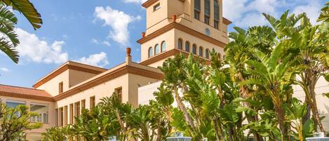 A hotel with a large building featuring a clock tower, surrounded by lush greenery and white tables set up for outdoor dining.