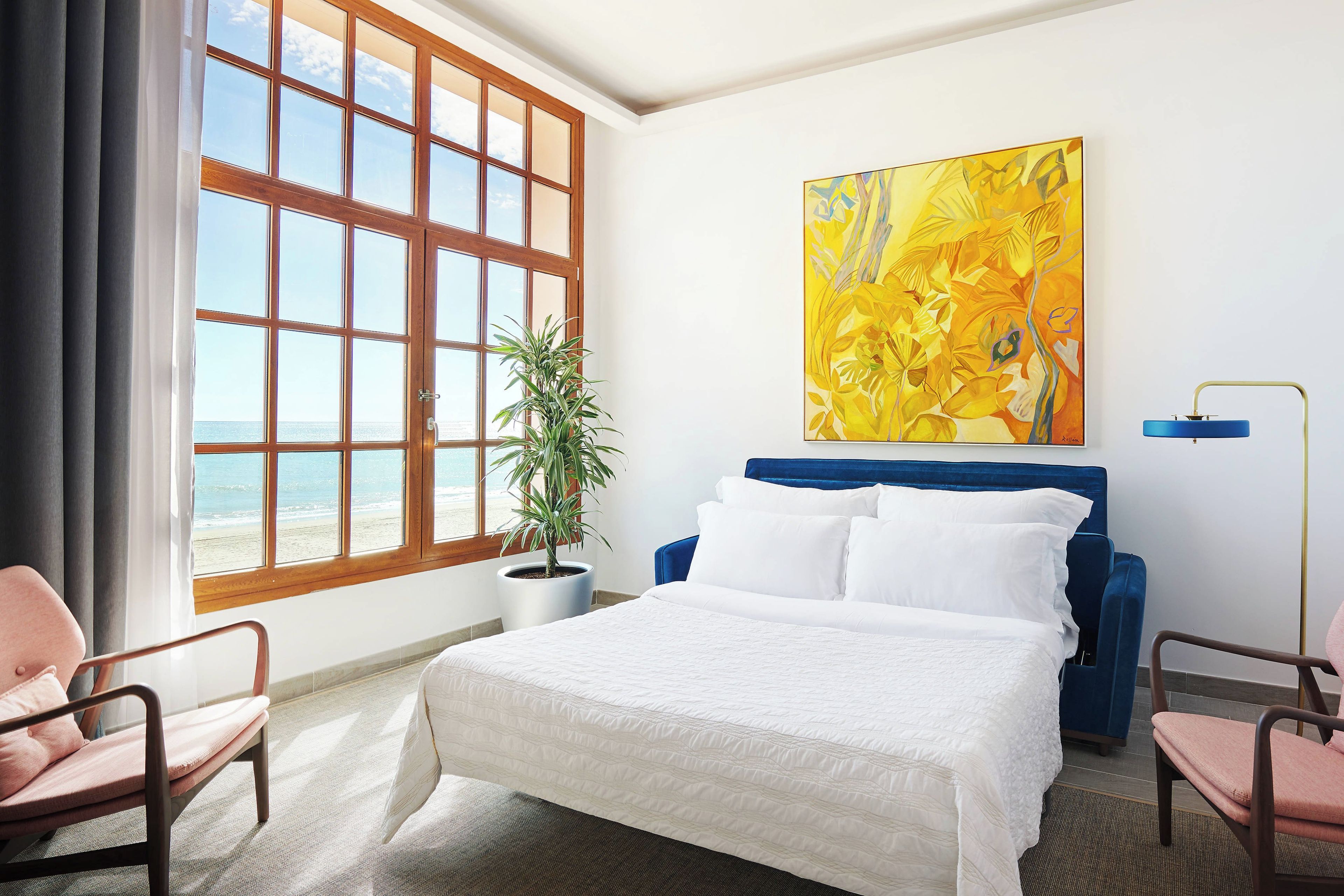 Een hotelkamer met een groot raam, een bed, een potplant en een schilderij aan de muur.