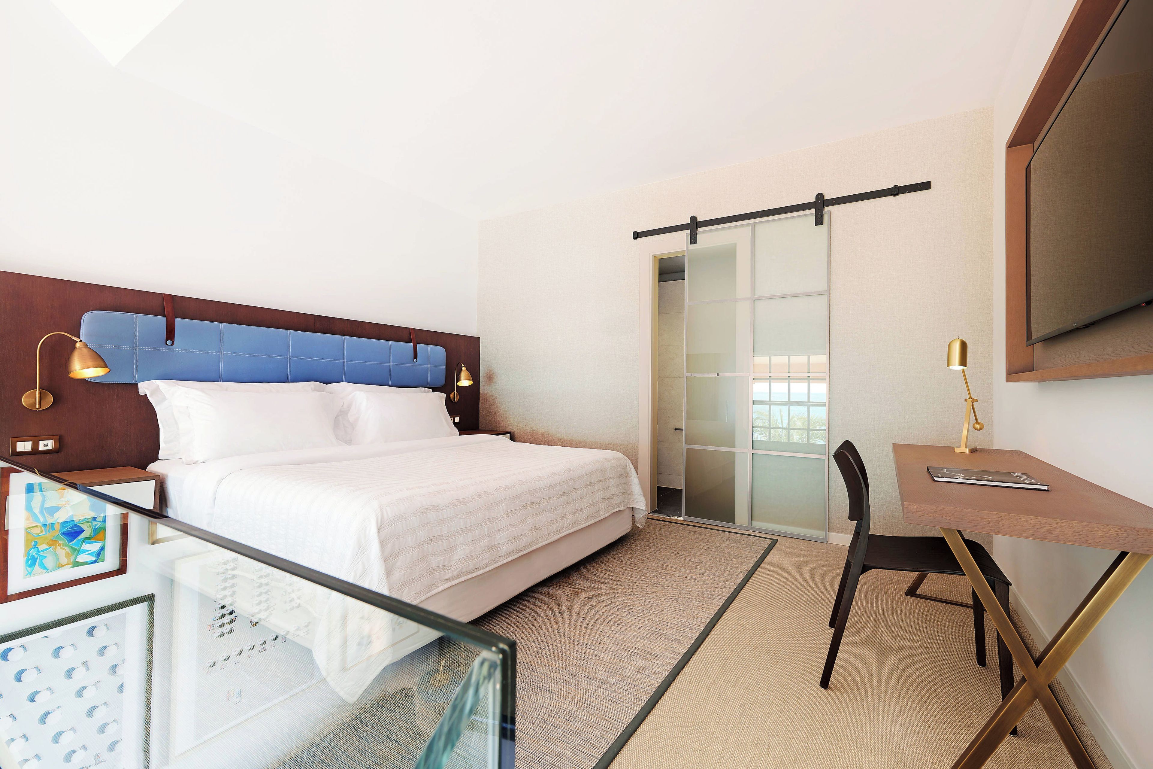 Quarto de hotel com cama, escrivaninha, cadeira, televisão e porta de correr.