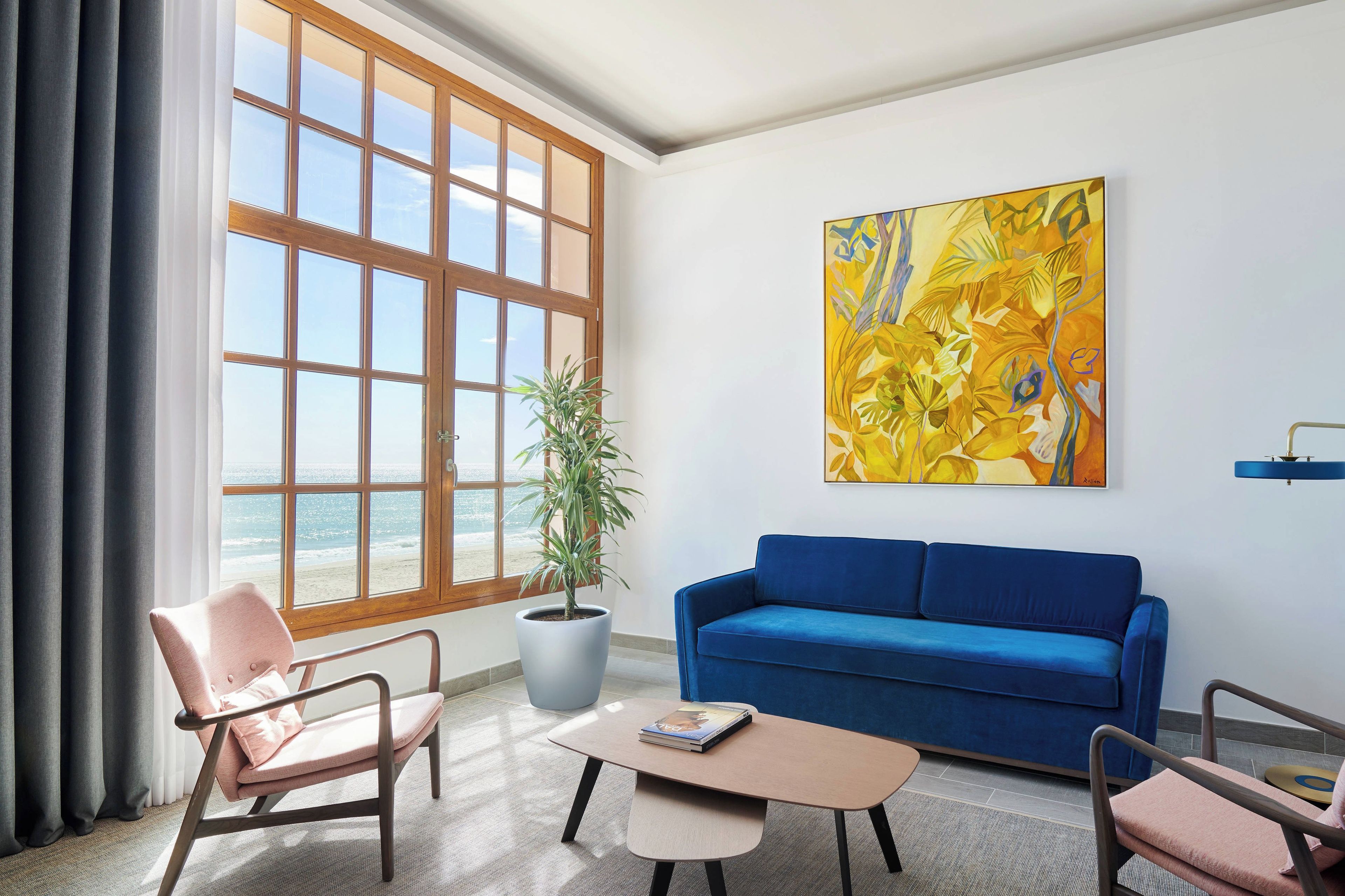 Uma sala de estar com uma janela ampla, um sofá azul, uma poltrona rosa, uma mesa de centro e um vaso de planta.