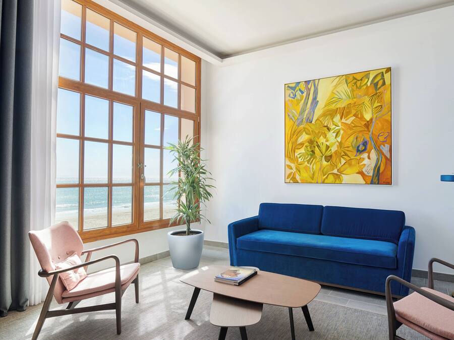 Uma sala de estar com uma janela ampla, um sofá azul, uma poltrona rosa, uma mesa de centro e um vaso de planta.