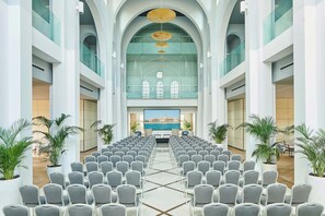 A conference room with rows of chairs facing a large screen, glass walls, and potted plants. - Le Meridien Ra Beach Hotel & Spa (El Vendrell)