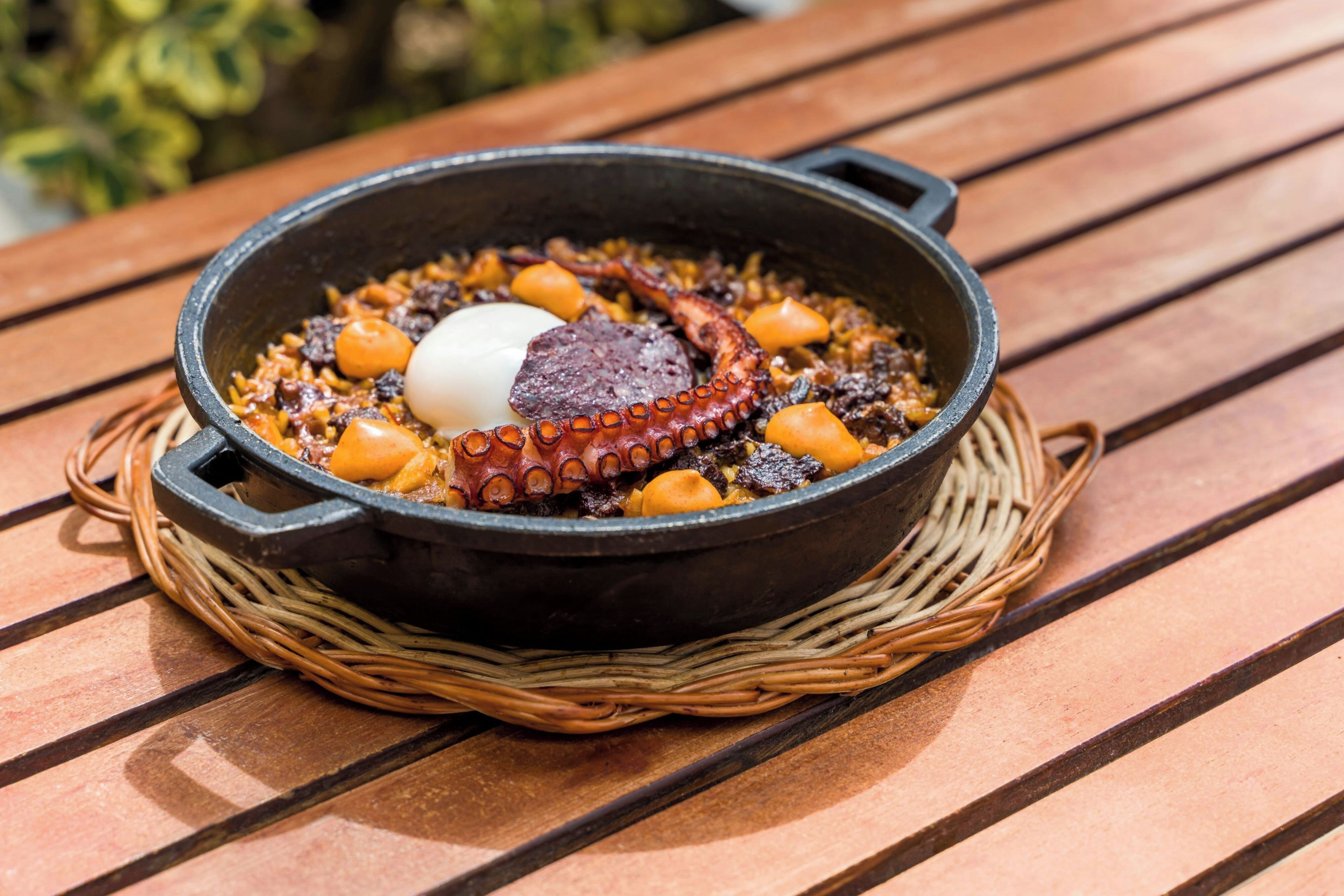 A black cast iron skillet with octopus and a boiled egg on a wooden table.