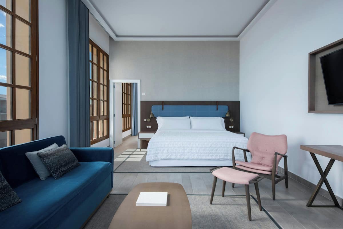A hotel room with a bed, a blue sofa, a pink chair, a small table, and a TV mounted on the wall.