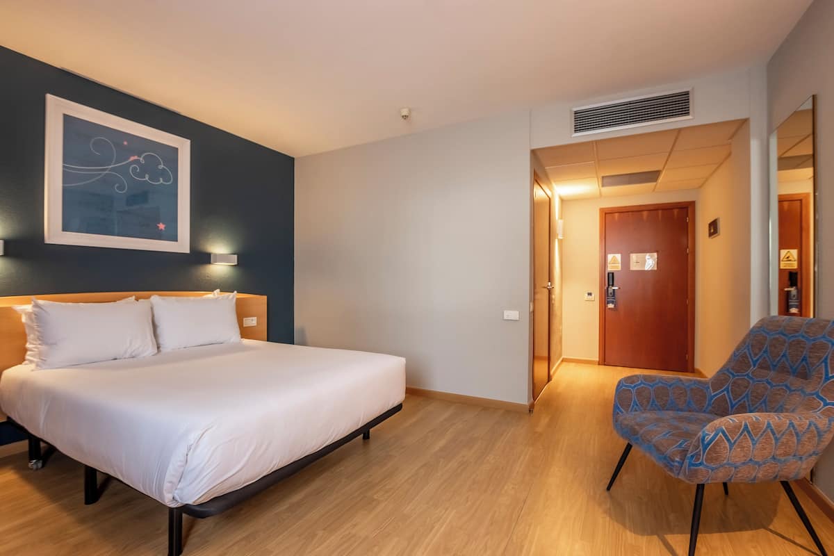 A hotel room with a bed, a chair, a framed artwork on the wall, and a door leading to another room.