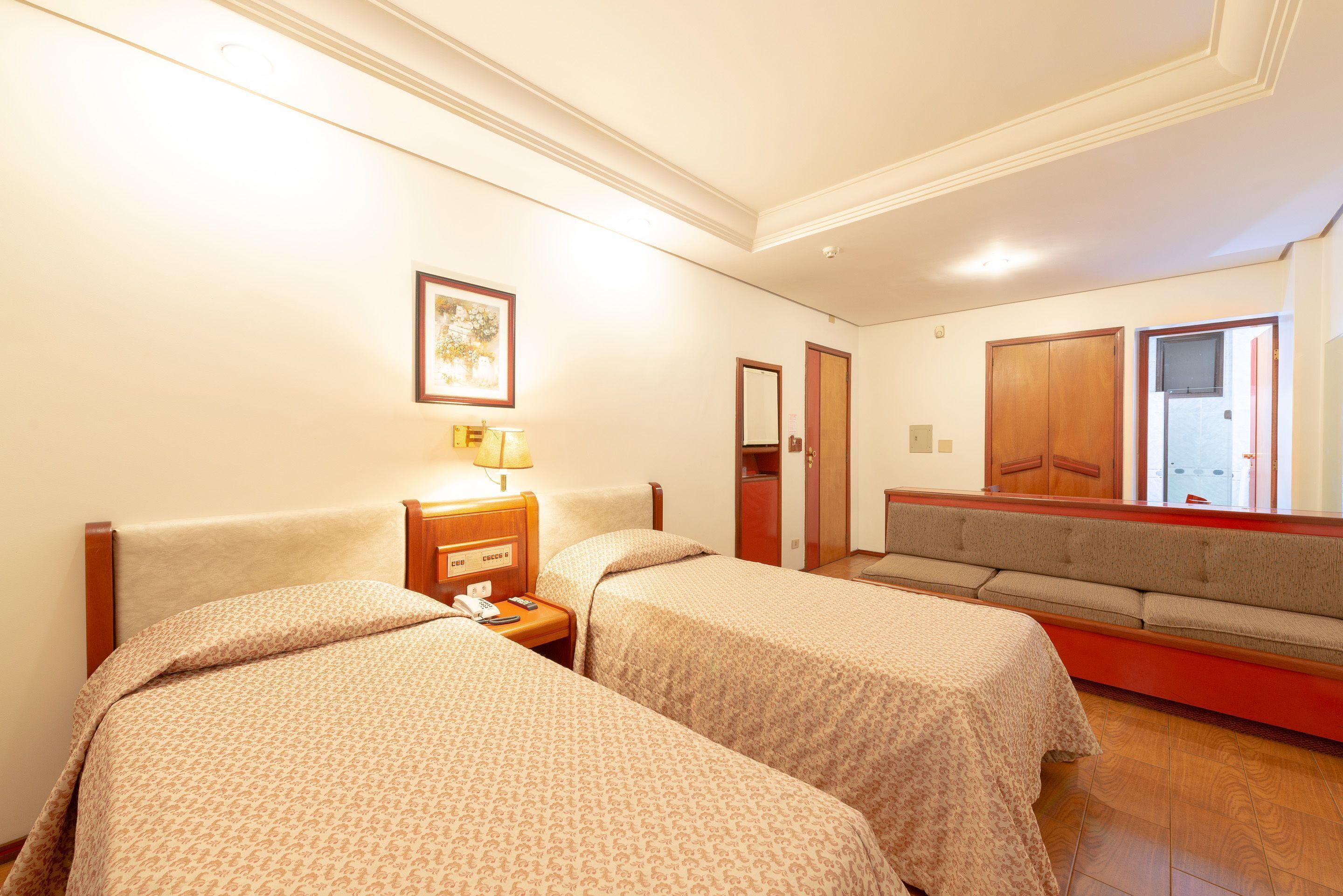 A hotel room with two beds, a sofa, a bedside table with a lamp, a framed picture on the wall, and a mirror.
