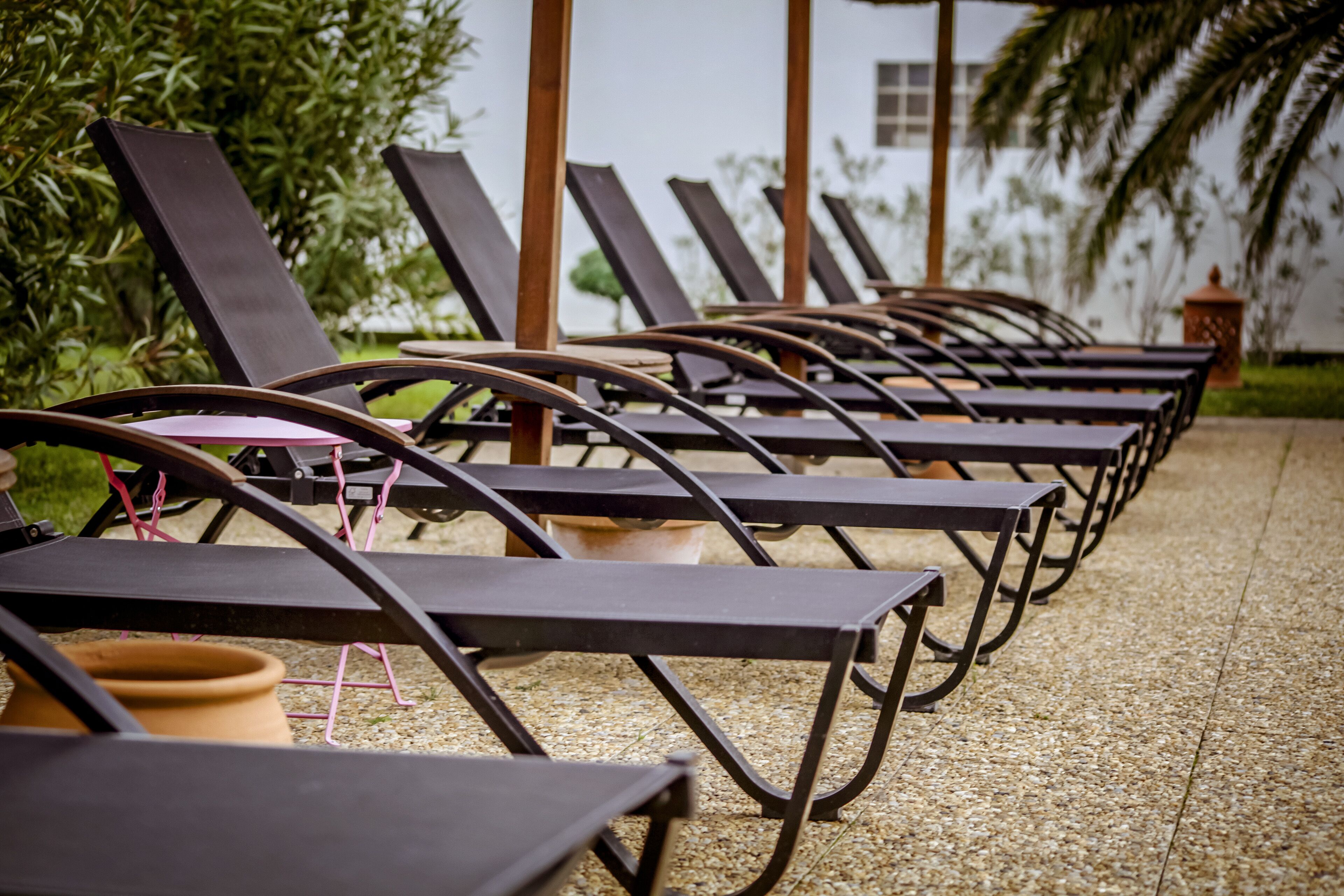 A row of sun loungers with metal frames and cushions, arranged on a gravel surface with greenery in the background.